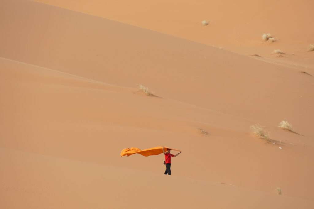 Person walking barefoot on Sahara dunes during a 5-day Fes to Marrakech desert tour, holding an orange scarf in the wind
